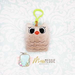 Op de afbeelding: Een beige pluche uil sleutelhanger met zwarte ogen, een rode snavel en een groene clip. De uil heeft geborduurde details en een klein bruin touw. De afbeelding bevat ook een kleine borduurring en de tekst "Miss Meggie Designs."