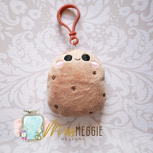 May include: A plush, light brown potato-shaped keychain with embroidered details. It features a smiling face with black eyes and pink cheeks, and brown spots. It has a brown clip attached to the top. The text "Miss Meggie Designs" is visible.