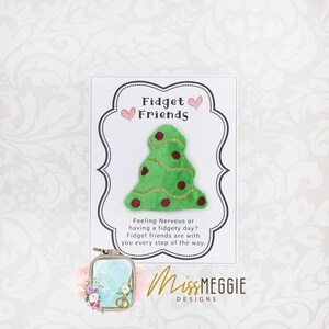 May include: A green Christmas tree-shaped fidget toy with red accents and gold trim is featured on a card that reads "Fidget Friends." The card also includes the text "Feeling Nervous or having a fidgety day? Fidget friends are with you every step of the way."