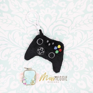 May include: A black plush video game controller keychain with a white loop. The controller has embroidered details in gray, white, and colorful buttons. The text "MissMEGGIE DESIGNS" is in gold script.