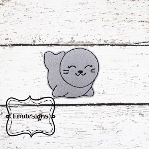May include: A gray embroidered seal with a happy expression, lying on its belly with its flippers outstretched. The seal is on a white wooden background.
