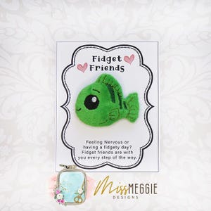 May include: A green plush fish with black eyes and a black outline, displayed on a white card with the text "Fidget Friends." The card also includes the text "Feeling Nervous or having a fidgety day? Fidget friends are with you every step of the way."