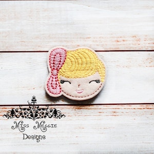 May include: Embroidered felt applique of a girl's face with blonde hair and a pink bow. The face has a smiling expression with closed eyes.