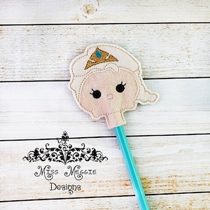 May include: A felt pencil topper with a cartoon character wearing a crown. The character has black eyes, a pink nose, and blonde hair. The pencil topper is attached to a turquoise pencil.