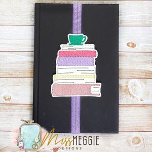 May include: A black journal with a purple ribbon bookmark. A stack of colorful books with a green teacup on top is stitched onto the cover. The journal has the words "Miss Meggie Designs" in cursive.