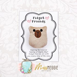 May include: A beige plush bear with a brown embroidered nose and black eyes, featured on a card with the text "Fidget Friends." The card also includes the text "Feeling Nervous or having a fidgety day?" and the brand name "MissMEGGIE DESIGNS."