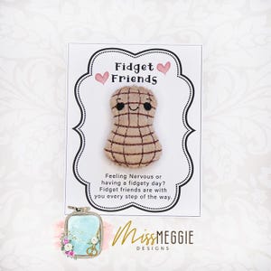 Puede incluir: Un juguete antiestrés beige con forma de cacahuete, con un patrón de cuadrícula y una cara sonriente. Se presenta en una tarjeta blanca con el texto "Fidget Friends" y un borde decorativo. El texto dice "Feeling Nervous or having a fidgety day?"