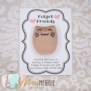 May include: A white card with a black border and pink hearts reads "Fidget Friends". A brown plush cat with a pink nose and black whiskers is attached to the card. The card also reads "Feeling Nervous or having a fidgety day? Fidget Friends are with you every step of the day." The bottom of the card reads "Miss Meggie Designs".