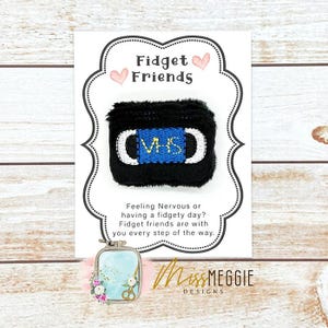 Puede incluir: Un juguete antiestrés en forma de cinta VHS negra con una etiqueta azul y detalles blancos, presentado en una tarjeta con el texto "Fidget Friends". La tarjeta también presenta un pequeño dije con detalles florales y de tijeras.