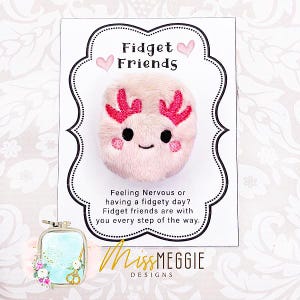 Puede incluir: Un juguete fidget de axolotl rosa con branquias rojas y ojos negros, presentado en una tarjeta con el texto "Fidget Friends". La tarjeta también incluye el texto "Feeling Nervous or having a fidgety day?" y el nombre del diseñador.