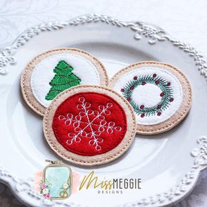 May include: Three embroidered Christmas coasters. One coaster features a green Christmas tree, another a red snowflake, and the third a green wreath with red berries. The coasters are round with a beige border and are on a white plate.