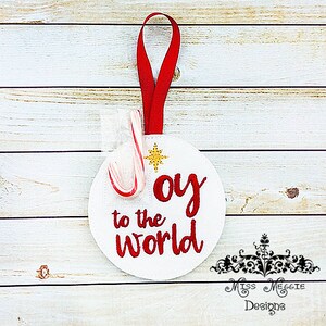 Könnte beinhalten: Runder weißer Anhänger mit roter Stickerei, auf dem "Joy to the world" steht. Ein Zuckerstange ist mit einem roten Band an dem Anhänger befestigt.