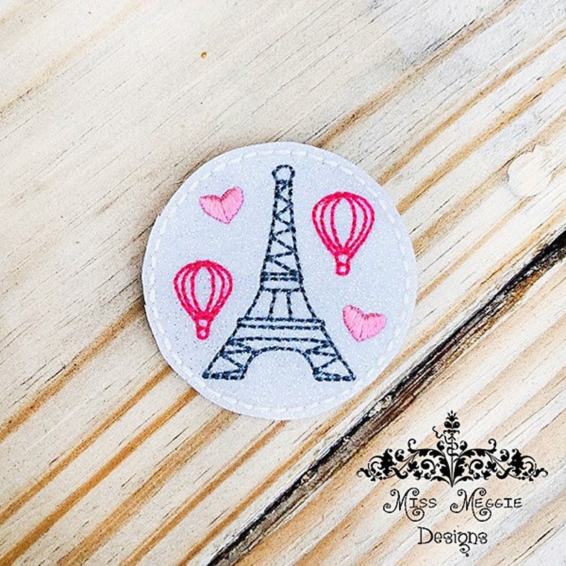 May include: A white felt circle with an embroidered design of the Eiffel Tower, two hot air balloons, and three pink hearts.