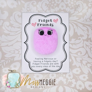 May include: Pink fuzzy fidget toy with a smiling face and eyelashes. The toy is on a white card with the text "Fidget Friends" and "Feeling Nervous or having a fidgety day? Fidget Friends are with you every step of the day". The card also has the text "MissMEGGIE DESIGNS".