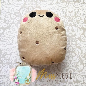 Puede incluir: Un juguete de peluche de patata marrón con una cara sonriente, mejillas rosadas y ojos bordados. La patata tiene una superficie texturizada y está diseñada para parecer una patata real.