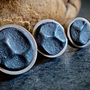 May include: Three circular stud earrings with a dark grey, textured surface and silver-coloured rims. The earrings are arranged in a row, with the largest in the foreground. The background is a dark, textured surface.