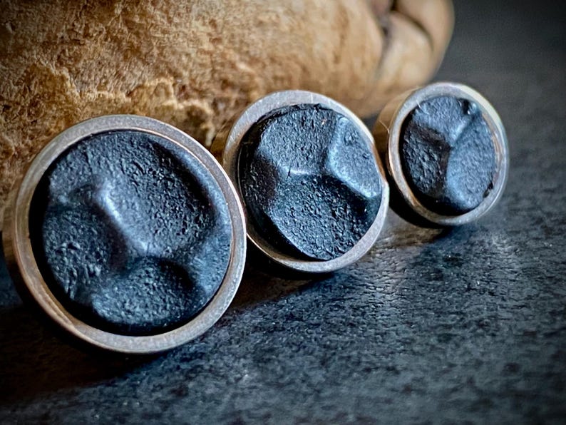 May include: Three circular stud earrings with a dark grey, textured surface and silver-coloured rims. The earrings are arranged in a row, with the largest in the foreground. The background is a dark, textured surface.