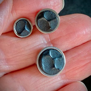 May include: Three silver-rimmed, dark grey metal buttons of varying sizes. Each button features a textured, geometric design within the circular frame. The buttons are displayed against a neutral background.