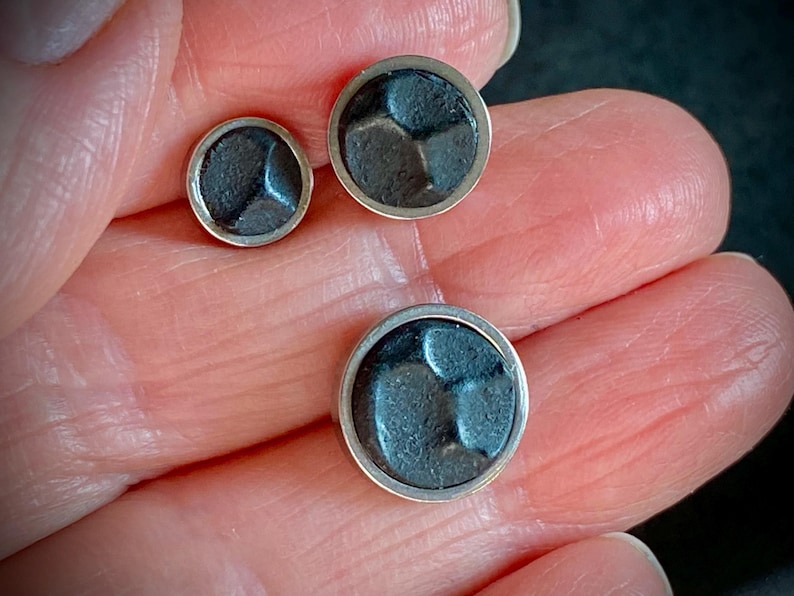 May include: Three silver-rimmed, dark grey metal buttons of varying sizes. Each button features a textured, geometric design within the circular frame. The buttons are displayed against a neutral background.