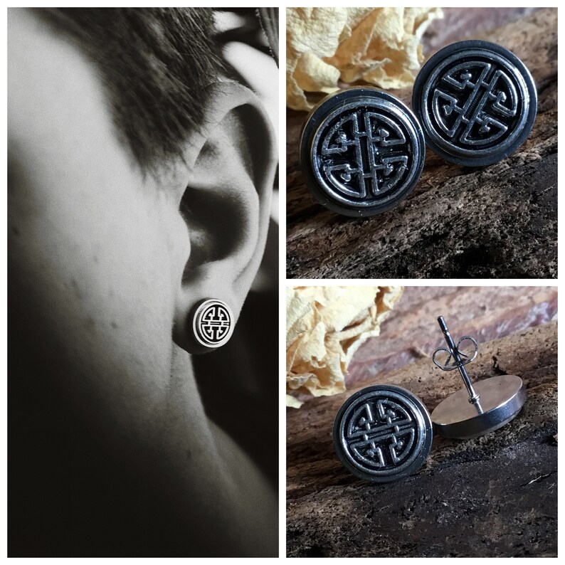 May include: A pair of silver stud earrings with a black circular design. The design features a stylized knot or symbol. The earrings are shown on a wooden surface and on a person's ear.