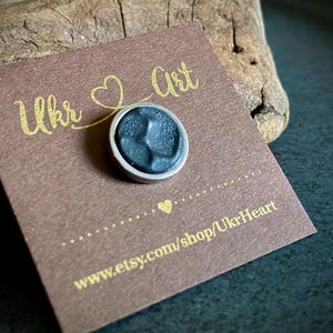May include: A small, round, dark grey pin with a silver border, on a brown card. The card has gold text reading "Ukr Art" and "www.etsy.com/shop/UkrHeart". The pin is on a dark surface.