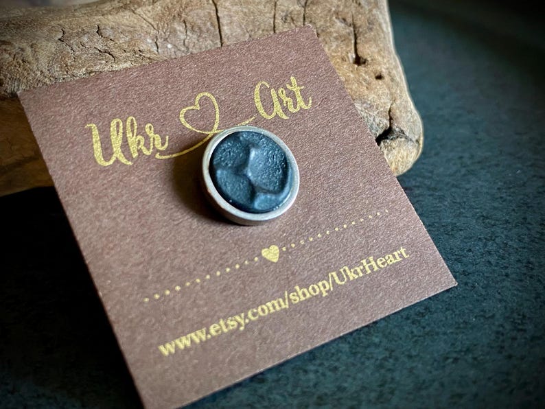 May include: A small, round, dark grey pin with a silver border, on a brown card. The card has gold text reading "Ukr Art" and "www.etsy.com/shop/UkrHeart". The pin is on a dark surface.
