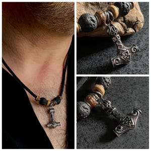 Puede incluir: Un collar de cordón de cuero negro con un colgante de Mjolnir plateado y cuentas de lava negra. El colgante tiene un diseño detallado con un patrón de nudos.