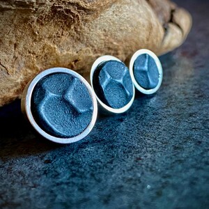 May include: Three dark blue stud earrings with a silver border. The earrings have a textured surface and a geometric design. They are arranged in a row, with a wooden element in the background.