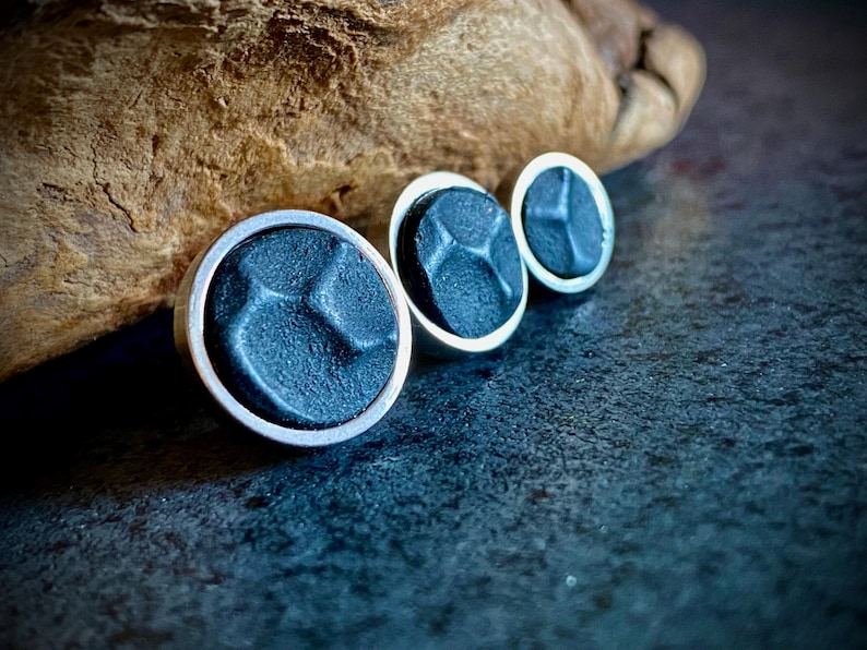 May include: Three dark blue stud earrings with a silver border. The earrings have a textured surface and a geometric design. They are arranged in a row, with a wooden element in the background.
