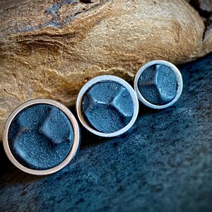 May include: Three round, dark grey metal buttons with a textured surface. The largest button has a copper-coloured rim, while the other two have silver-coloured rims. The buttons are arranged on a dark surface with a wooden background.