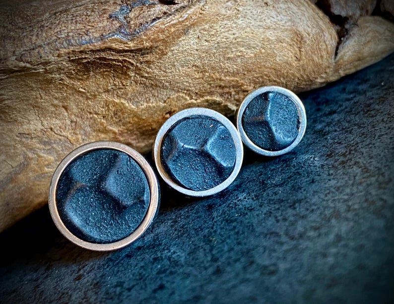 May include: Three round, dark grey metal buttons with a textured surface. The largest button has a copper-coloured rim, while the other two have silver-coloured rims. The buttons are arranged on a dark surface with a wooden background.