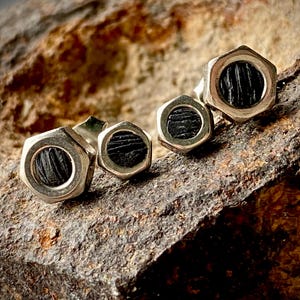 Puede incluir: Un par de pendientes de tuerca hexagonales plateados con un centro negro texturizado. Los pendientes se muestran sobre una superficie de piedra marrón rugosa, creando un contraste.