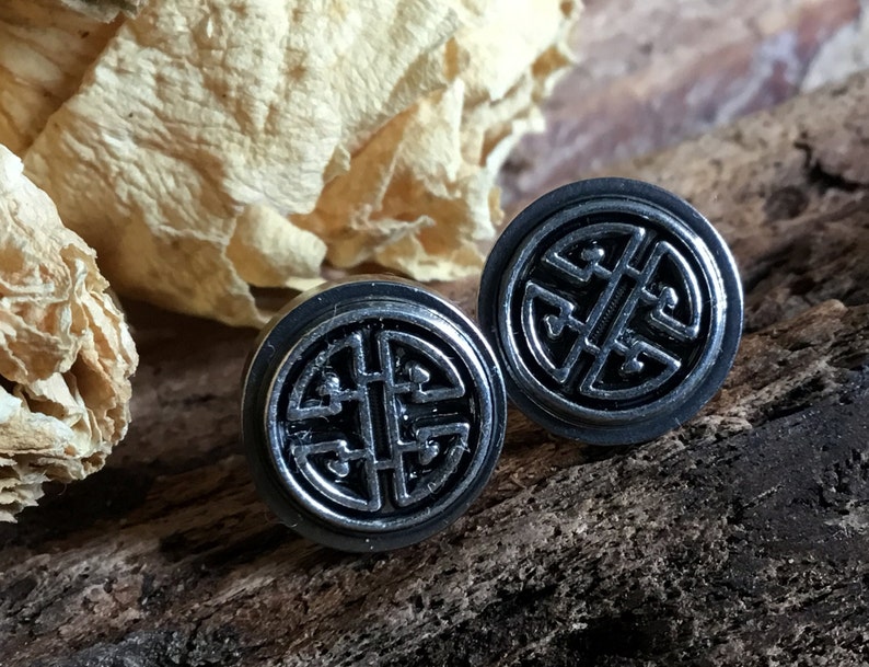 May include: A pair of silver stud earrings with a black, intricate knot design. The earrings are round and have a polished finish.