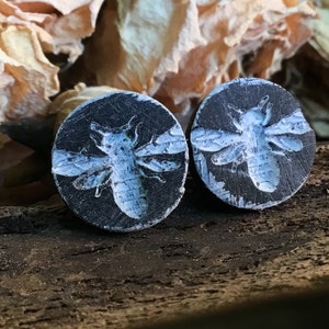 May include: Two round, black earrings with a light blue bee design. The bee is centered on the earring, with detailed wings and body. The earrings are set against a rustic, wooden background with dried flowers.