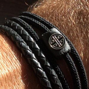 Könnte beinhalten: Schwarzes Lederarmband mit mehreren geflochtenen Strängen. Das Armband hat einen silberfarbenen, kreisförmigen Verschluss mit einem komplizierten Design. Das Leder scheint hochwertig zu sein und das Armband ist für das Handgelenk gedacht.