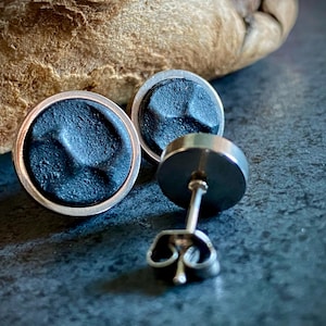 May include: Close-up of two round, dark grey stud earrings with a textured surface and silver-coloured trim. One earring post is visible. The earrings are on a dark grey surface with a blurred wooden background.