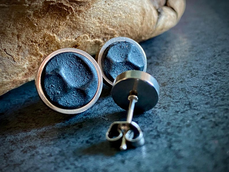 May include: Close-up of two round, dark grey stud earrings with a textured surface and silver-coloured trim. One earring post is visible. The earrings are on a dark grey surface with a blurred wooden background.