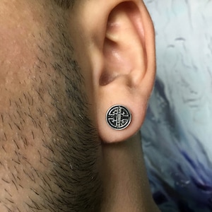 May include: A close-up of a person's ear with a silver ear gauge featuring a black circular design. The design is a stylized geometric pattern.