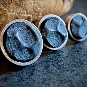 May include: Close-up of three dark grey metal cufflinks with a textured, geometric design. Each cufflink has a circular silver-coloured border. The cufflinks are arranged in a row, with a blurred background.