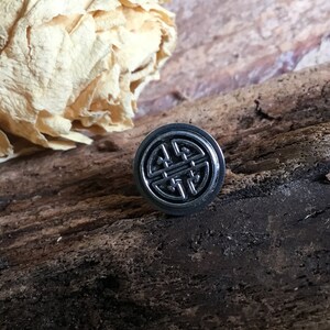 May include: A silver-colored metal ring with a black, intricate design. The ring is on a wooden surface.