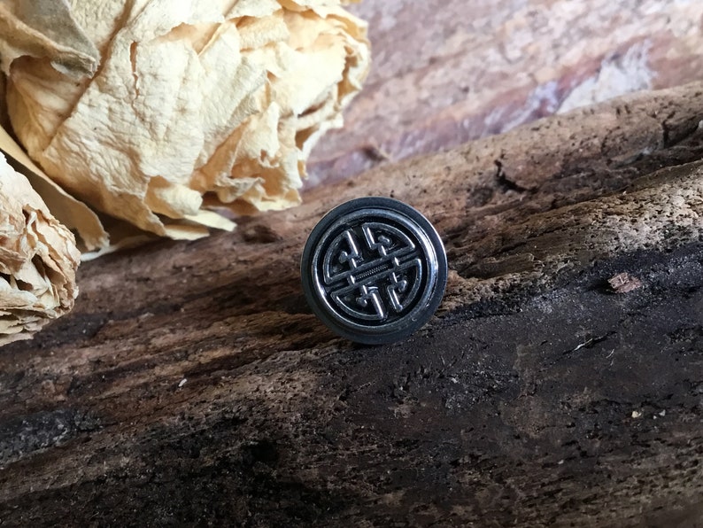 May include: A silver-colored metal ring with a black, intricate design. The ring is on a wooden surface.