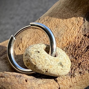 Könnte beinhalten: Ein silberner Creolen-Ohrring mit einem kleinen, beige-farbenen, donut-förmigen Steinanhänger. Der Ohrring ist aus glänzendem Metall gefertigt und der Stein hat eine raue Textur. Der Ohrring wird auf einem Holzstück präsentiert.