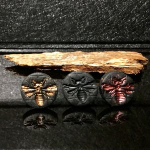 May include: Three round, black buttons with embossed bee designs. One button has a gold bee, one has a black bee, and the third has a red bee. The buttons are displayed on a reflective black surface.