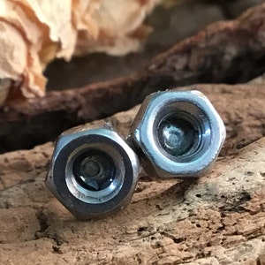 May include: Two silver metal hexagonal nuts with a small hole in the centre of each. The nuts are lying on a piece of wood.