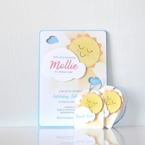May include: A blue and yellow birthday invitation with a sun and cloud design. The invitation says "Our little sunshine Mollie is turning one." It also includes the text "Saturday, February" and "RSVP to MEGA".