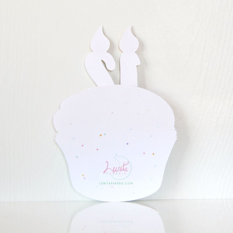 May include: White cupcake card with two candles on top. The card has a light blue background with white sprinkles and the text "Lunita Paperie" in a light blue font.