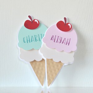 May include: Two ice cream cone shaped place cards with red cherries on top. The ice cream is in layers of white, pink, and light blue. The place cards are labeled "Charlie" and "Aliyah".