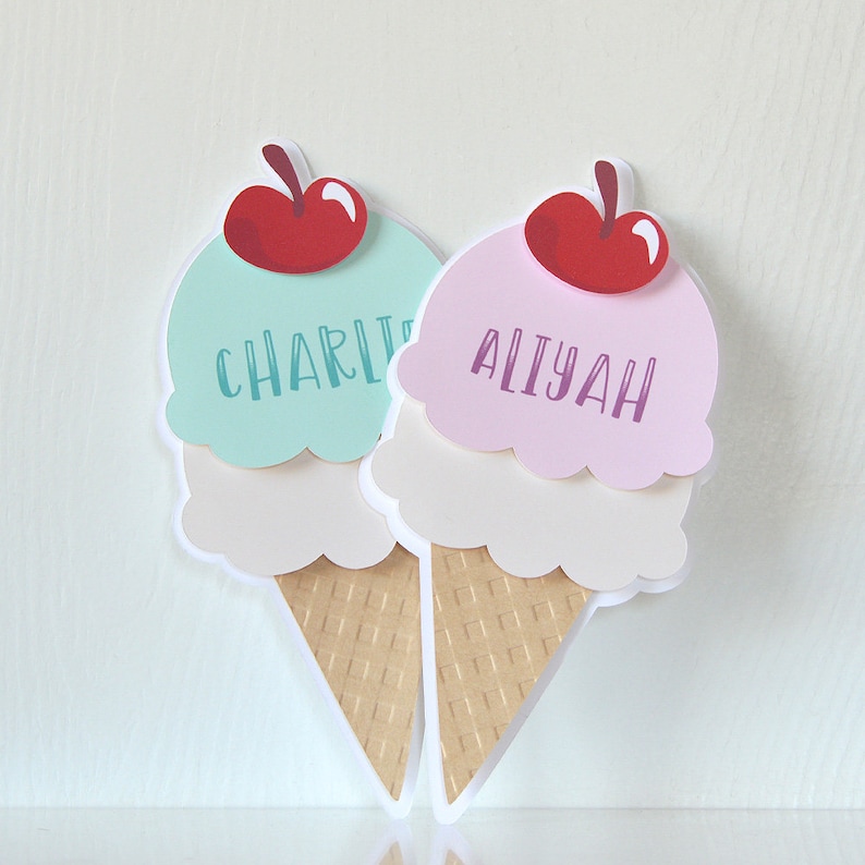 May include: Two ice cream cone shaped place cards with red cherries on top. The ice cream is in layers of white, pink, and light blue. The place cards are labeled "Charlie" and "Aliyah".