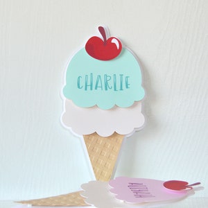 May include: Two ice cream cone shaped place cards with the names Charlie and Aliyah. The ice cream cones are made of paper and have a light blue, white, and brown color scheme. The ice cream cones have a cherry on top.