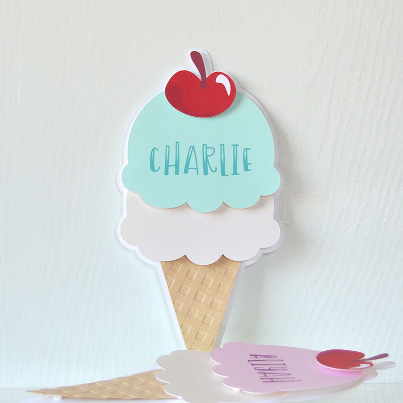 May include: Two ice cream cone shaped place cards with the names Charlie and Aliyah. The ice cream cones are made of paper and have a light blue, white, and brown color scheme. The ice cream cones have a cherry on top.